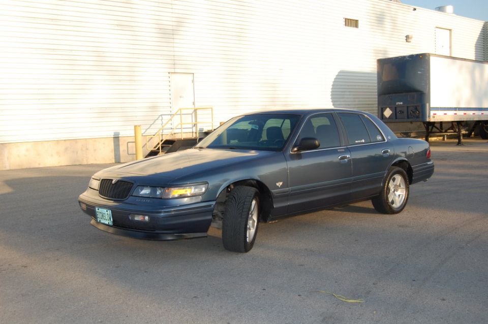 Blue-gray Mercury Grand Marquis sedan, front-left view, with alloy wheels in an industrial lot