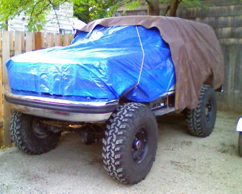 1993 Ford Bronco - An SUV, possibly a 1993 Ford Bronco, is covered by tarps, with only its tires and front bumper visible.