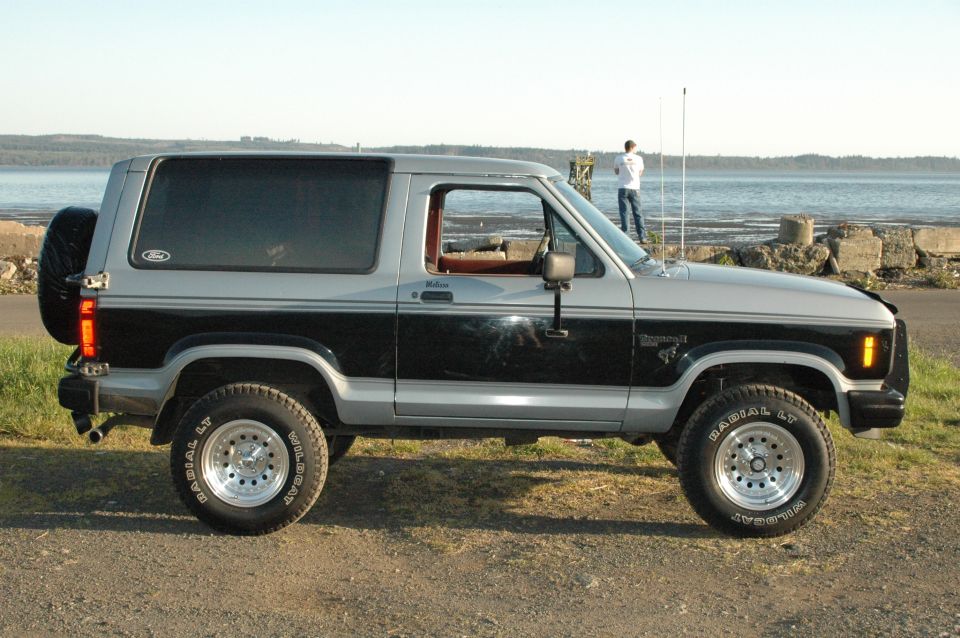 Black and silver Ford Bronco II SUV in side view, lifted with chrome wheels, parked by water.
