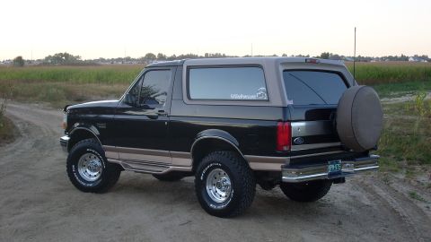 1996 Ford Bronco - This 1996 Ford Bronco is viewed from its rear three-quarter angle, parked on a dirt road with fields in the background.