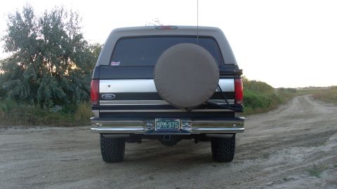 1996 Ford Bronco - The rear of a 1996 Ford Bronco is shown parked on a dirt road with vegetation in the background.