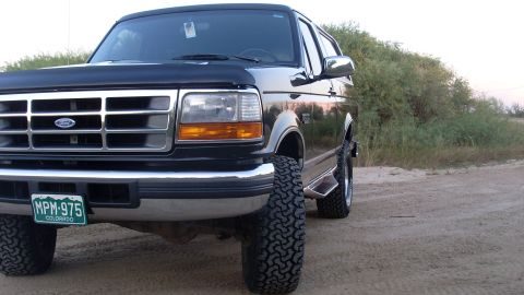 1996 Ford Bronco - A black Ford Bronco is parked on a dirt road with brush in the background.
