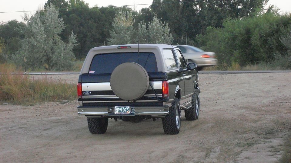1996 Ford Bronco parked on dirt road, spare tire on rear door. Another car is blurred in motion behind it