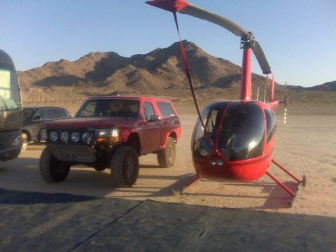 1984 Ford Bronco - Modified red Ford Bronco parked next to a red helicopter in a desert setting.