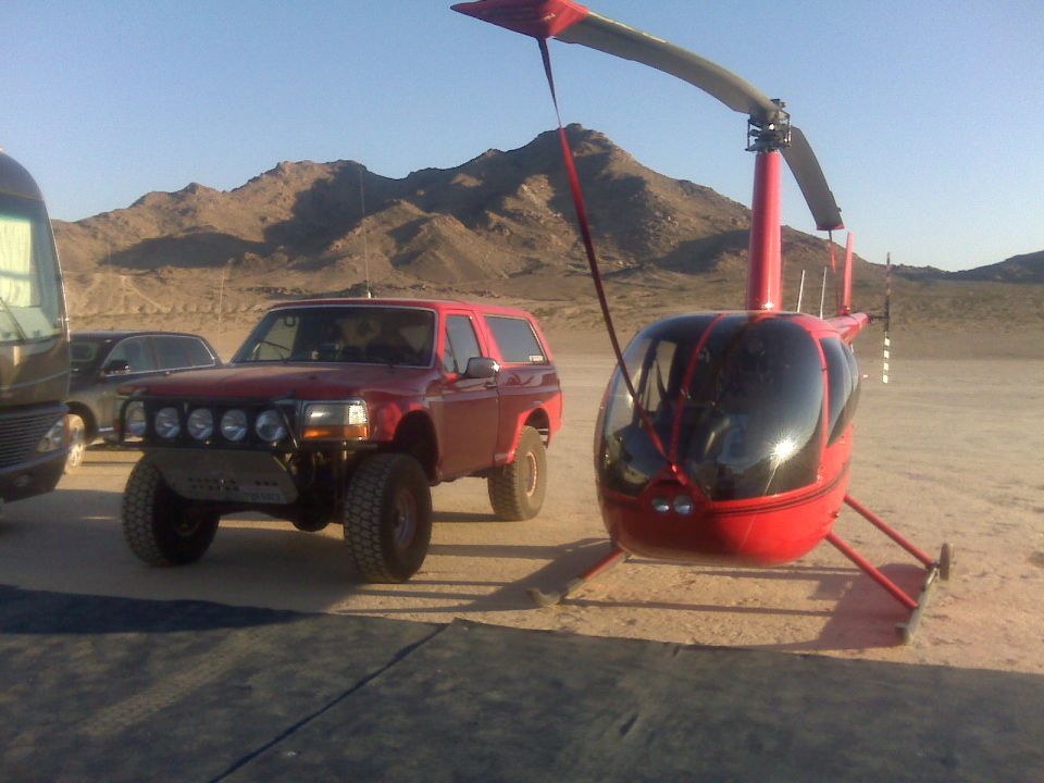 Red Ford Bronco with aftermarket bumper and lights next to a red helicopter in a desert landscape
