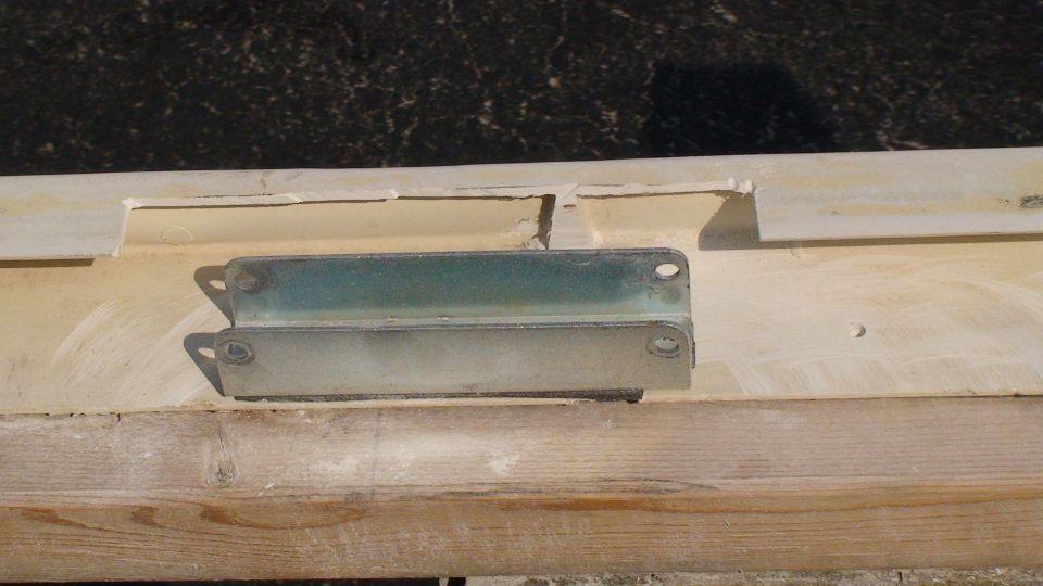 Zinc-plated four-hole steel bracket on a cream painted sheet metal channel resting on lumber