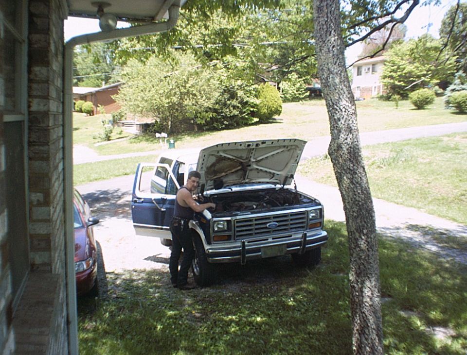 White and blue Ford Bronco with hood up in driveway, man working on engine