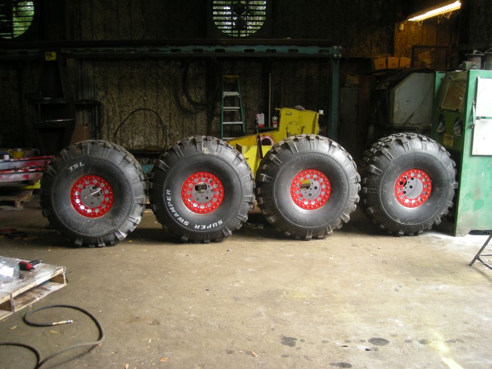 Four large off-road tires with red beadlock rims lined up on a concrete floor