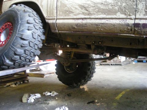 1979 Ford Bronco - The underside of a heavily modified off-road vehicle, covered in mud, is shown in a garage setting.