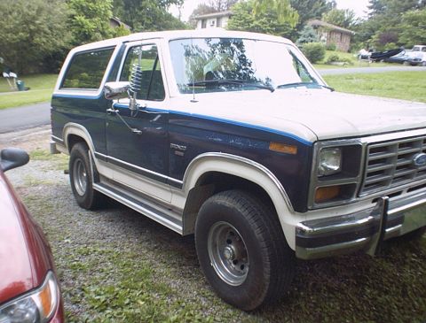1986 Ford Bronco - A two-tone blue and white Ford Bronco is parked on a gravel surface with trees and houses in the background.