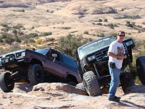 1956 Ford Bronco II - Two heavily modified off-road vehicles navigate a challenging rocky trail in a desert environment.