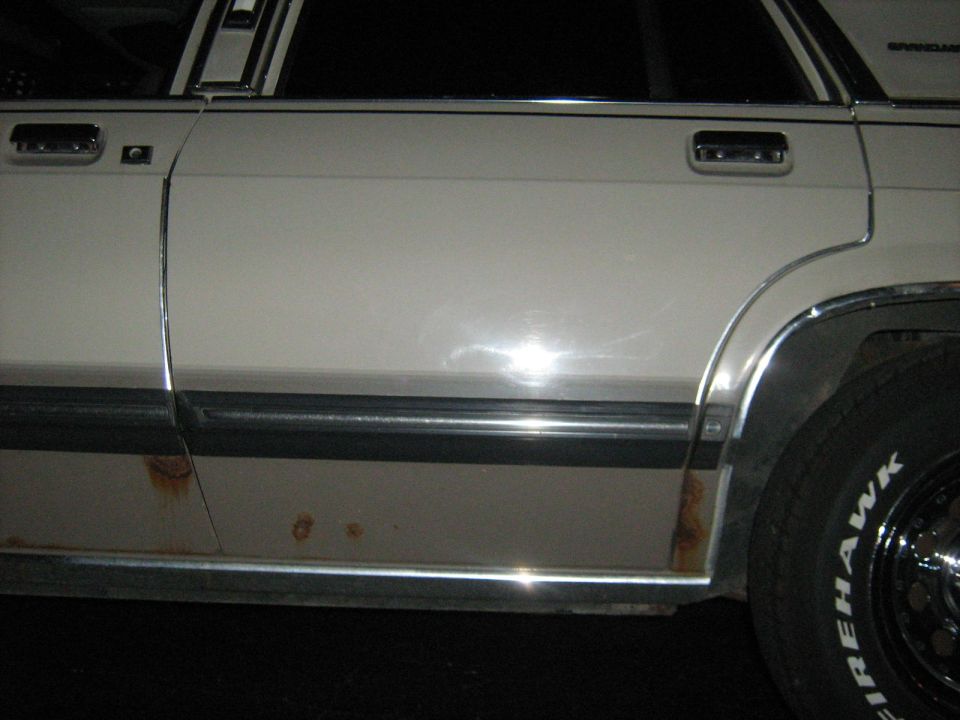 White 1988 Mercury Grand Marquis side view with rust spots on lower body and chrome trim