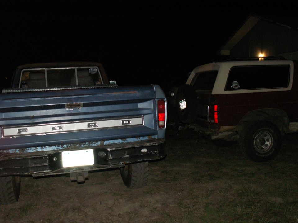 Rear view of a blue 1977 Ford F-250 pickup truck parked next to a red Ford Bronco at night.