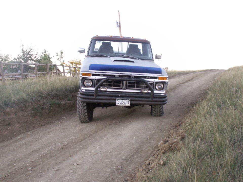 Lifted 1977 Ford F-250 pickup truck with blue hood accent and bull bar on a dirt road.