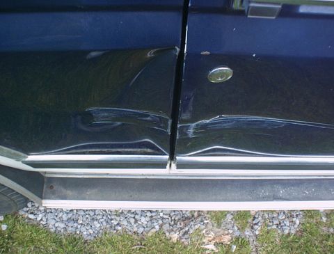1986 Ford Bronco - The side panel and door of a dark blue vehicle show significant damage and creasing.