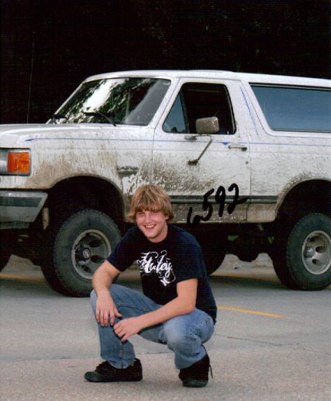 1987 Ford Bronco - A young man squats in front of a muddy white Ford Bronco. The Bronco has the number "592" written on its side.