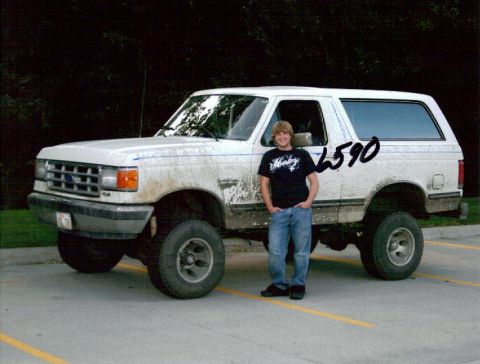 1987 Ford Bronco - A young man stands next to a dirty white 1987 Ford Bronco. The Bronco is a two-door model with a lifted suspension and large tires.