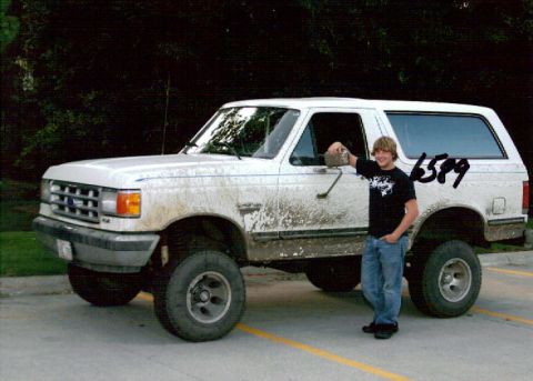 1987 Ford Bronco - A young man stands next to a dirty white 1987 Ford Bronco with aftermarket wheels. The vehicle is parked on a paved surface with yellow lin…