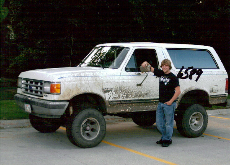 White 1987 Ford Bronco with mud splatters and aftermarket wheels, parked outside with a young man standing next to it.