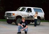 1987 Ford Bronco - A lifted white Ford Bronco is parked on asphalt with a young man squatting in front of it and a young woman standing behind it.