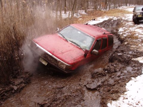 1989 Ford Bronco - A red SUV is stuck in deep mud, with the front tires submerged and mud splashing around the wheel well. Another off-road vehicle is visible…