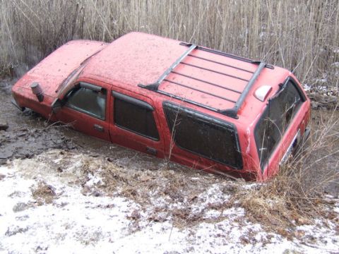1989 Ford Bronco - A red SUV is stuck in a muddy ditch with snow on the ground and tall dry grass surrounding it. The vehicle is partially submerged in the mu…