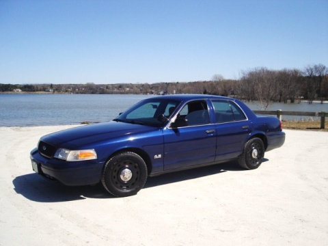 2003 Ford Crown Victoria - A dark blue 2003 Ford Crown Victoria is parked on a light-colored surface next to a body of water. The car is viewed from a 3/4 front angle…