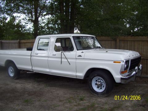 1978 Ford Bronco - A white four-door pickup truck is parked on a dirt lot in front of a wooden fence. The truck has chrome accents on the grille and bumper, a…