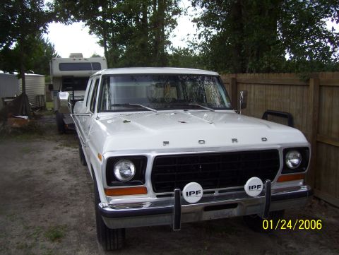 1978 Ford Bronco - A white 1970s Ford pickup truck is parked outdoors on a dirt lot, with a camper trailer visible behind it. The truck features a chrome bump…