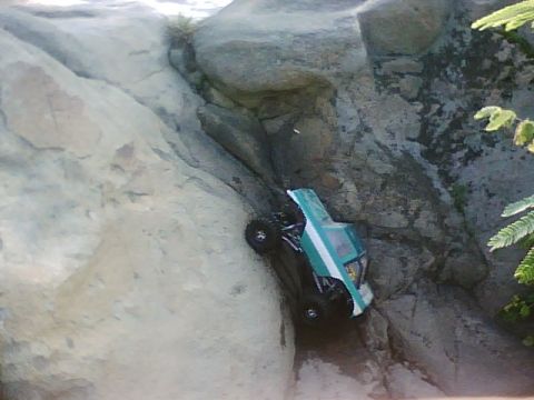 1996 Ford Bronco - A teal and white RC rock crawler truck is navigating a steep, rocky incline. The truck is positioned at an angle, with its wheels gripping…
