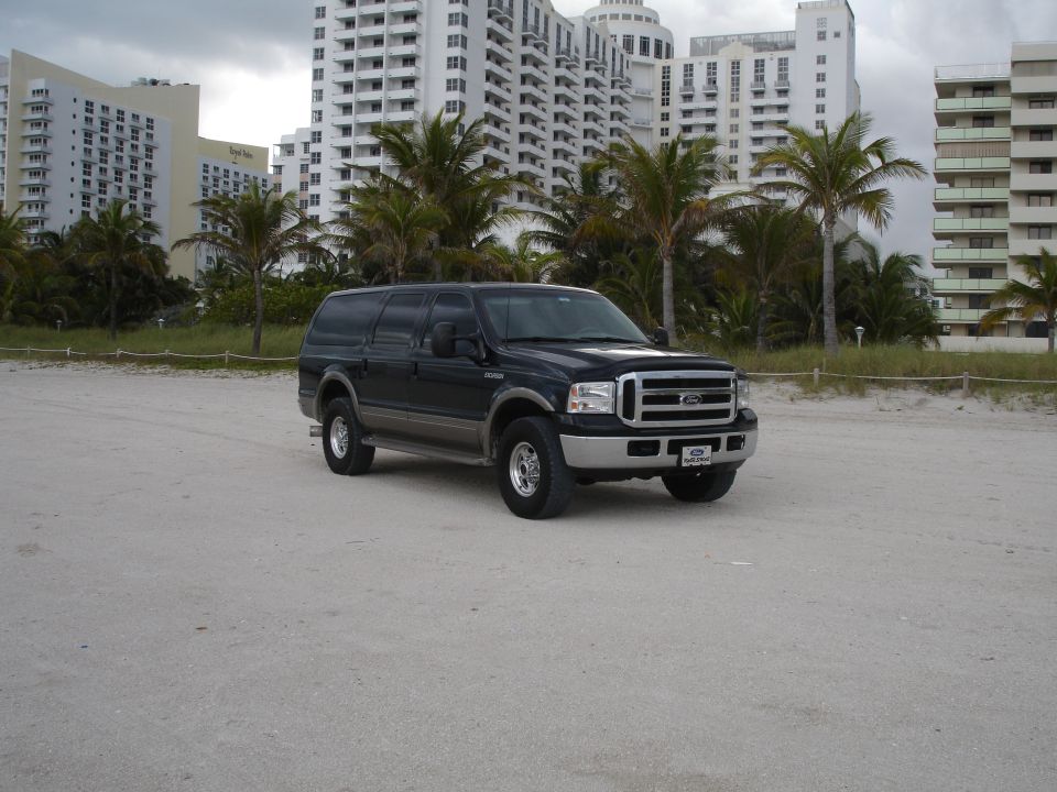 Dark Ford Excursion parked on a beach with buildings and palm trees in the background.