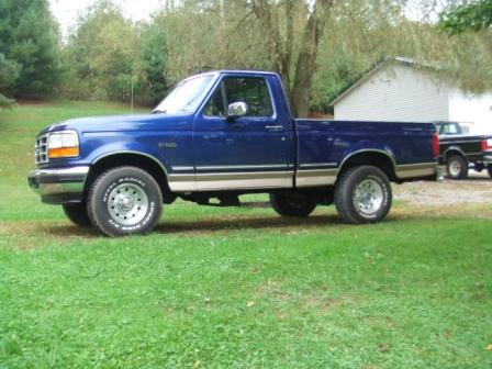 1996 Ford Bronco - A blue Ford pickup truck with a tan stripe is parked on a grassy lawn in front of a white building. The truck is shown from the side view,…