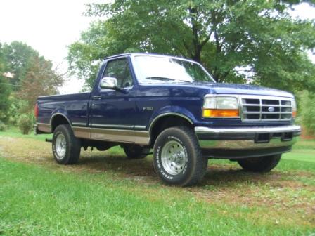 1996 Ford Bronco - A blue Ford F-150 pickup truck is parked on a grassy lawn with trees in the background. The truck is shown from a three-quarter front angle…