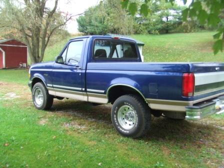 1996 Ford Bronco - A blue 1990s Ford pickup truck is parked on a grassy lawn next to a red barn. The truck is viewed from the rear quarter angle, showing its…