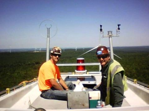 1996 Ford Bronco - Two men in hard hats and safety vests are sitting on the platform of a wind turbine, with a forest and other wind turbines visible in the b…