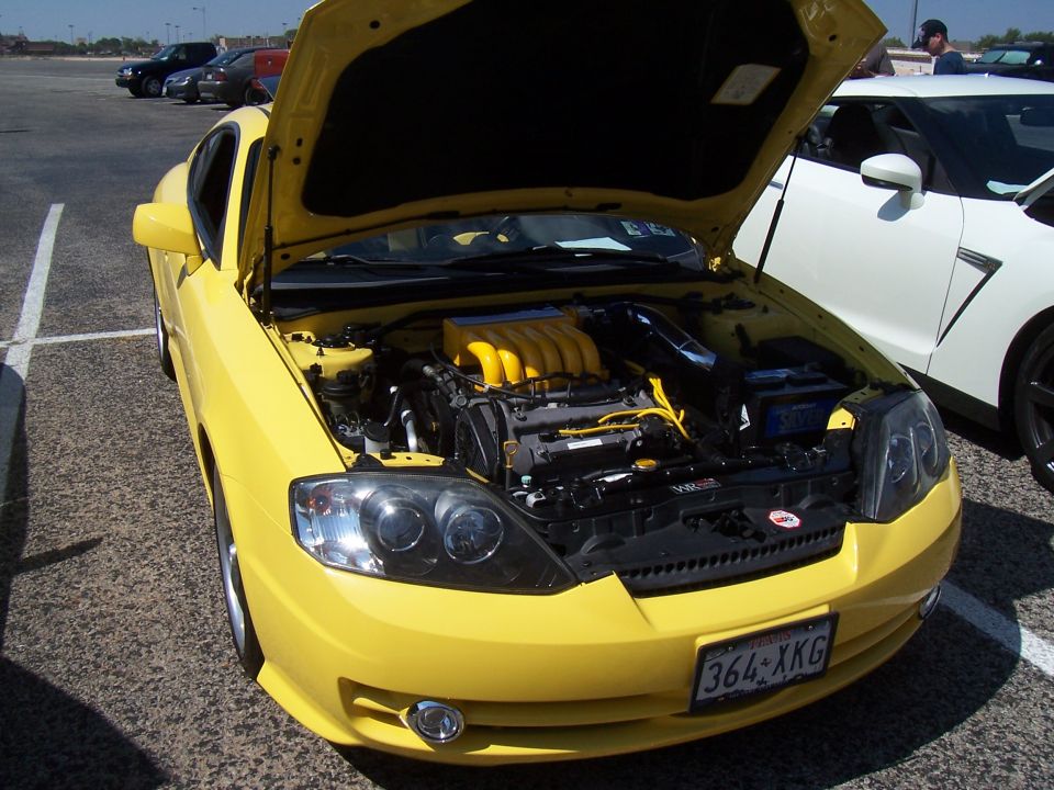 Yellow coupe with hood open, revealing a yellow-topped engine. License plate reads 364-XKG. A white sports car is parked to the right