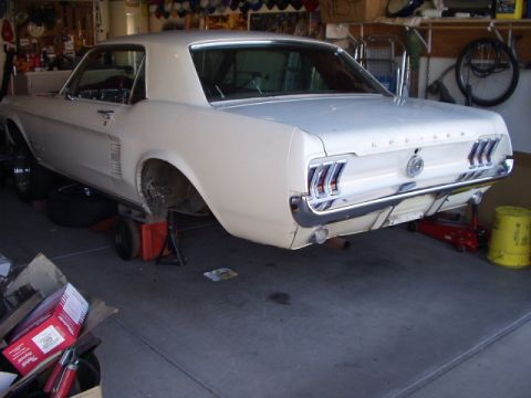 1967 Ford Mustang - A white 1967 Ford Mustang coupe is shown from the rear while undergoing maintenance in a garage.
