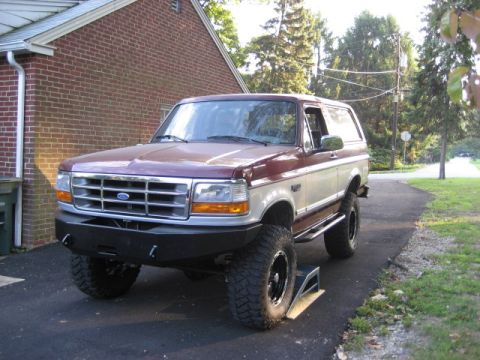 1996 Ford Bronco - A two-tone 1996 Ford Bronco is shown from a three-quarter front angle, parked on a paved surface next to a brick house.