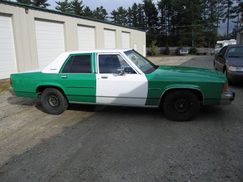1991 Ford LTD Crown Victoria - two-tone green and white full-size Ford sedan is parked in profile on gravel in front of metal storage buildings.