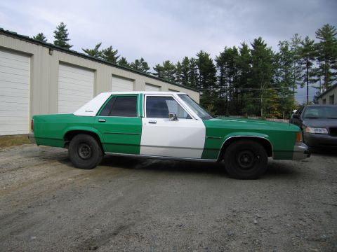 1991 Ford LTD Crown Victoria - green and white full-size Ford sedan sits in side profile on a gravel lot with storage units and pines behind it.