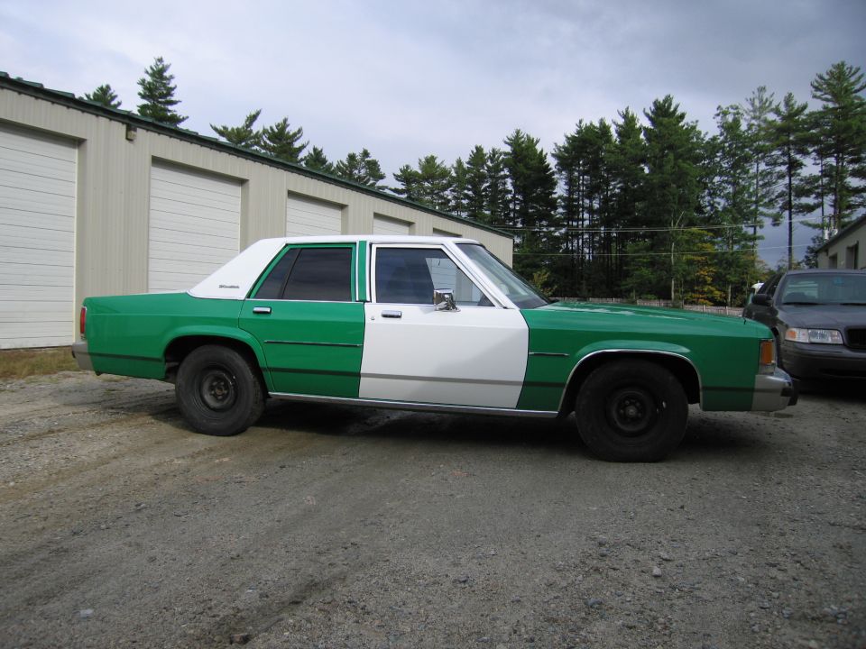 Green and white Ford sedan, driver-side profile, parked on gravel lot
