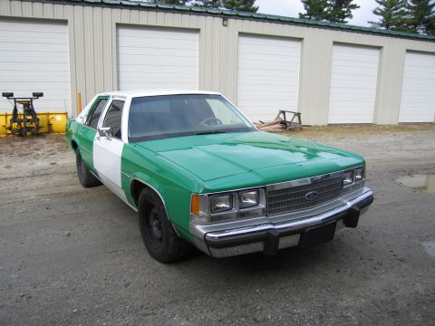 1991 Ford LTD Crown Victoria - Front three-quarter view of a green and white full-size Ford sedan, its chrome grille bearing the Ford emblem, parked on gravel.