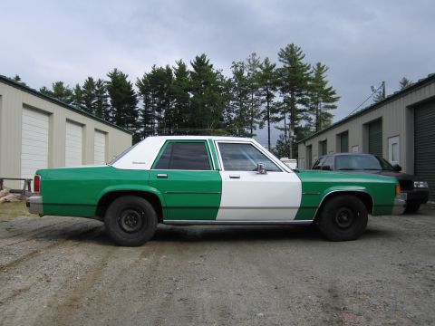 1991 Ford LTD Crown Victoria - Passenger-side profile of a green and white full-size Ford sedan parked on a gravel lot in front of metal buildings.