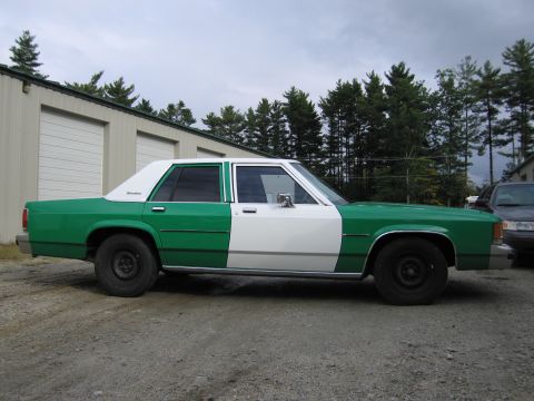 1991 Ford LTD Crown Victoria - Passenger-side profile of a green and white full-size Ford sedan parked on gravel with another vehicle and trees behind.