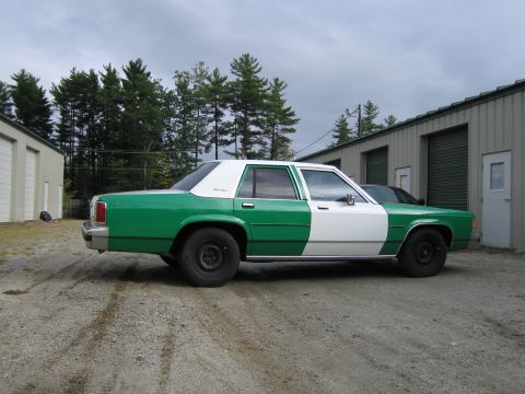 1991 Ford LTD Crown Victoria - Rear three-quarter passenger-side view of a green-and-white full-size Ford sedan on a gravel lot beside green metal garages.