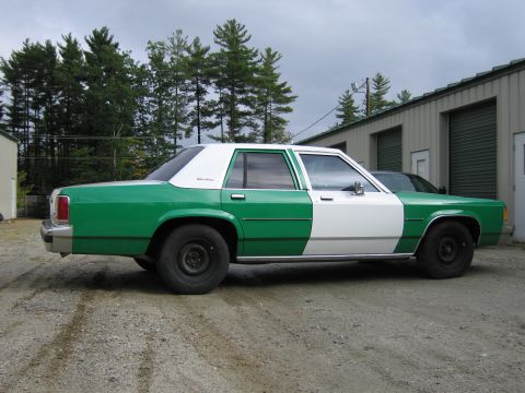 1991 Ford LTD Crown Victoria - Passenger-side profile of a green-and-white two-tone full-size Ford sedan parked on gravel in front of pine trees and a metal building.
