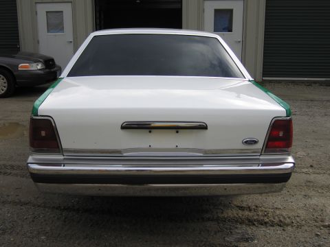 1991 Ford LTD Crown Victoria - Straight-on rear view of a white full-size Ford sedan with green trim, Ford oval and chrome bumper, parked before garage bays.