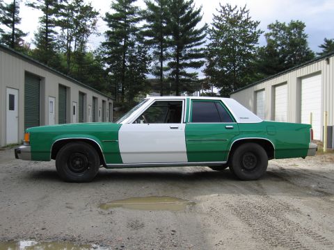 1991 Ford LTD Crown Victoria - Driver-side profile of a green-and-white two-tone full-size Ford sedan parked on a gravel lot in front of storage sheds and tall pines.
