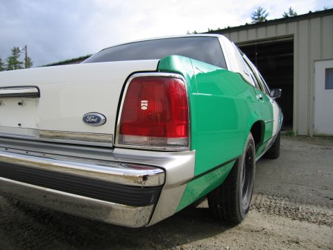 1991 Ford LTD Crown Victoria - Rear quarter close-up of a white-and-green full-size Ford sedan showing the Ford oval, taillight and chrome bumper near a garage.