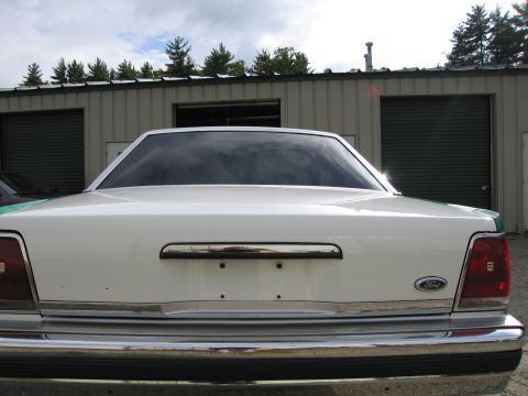 1991 Ford LTD Crown Victoria - Straight-on rear view of a white full-size Ford sedan with green edges, Ford oval and chrome bumper, parked under cloudy sky.